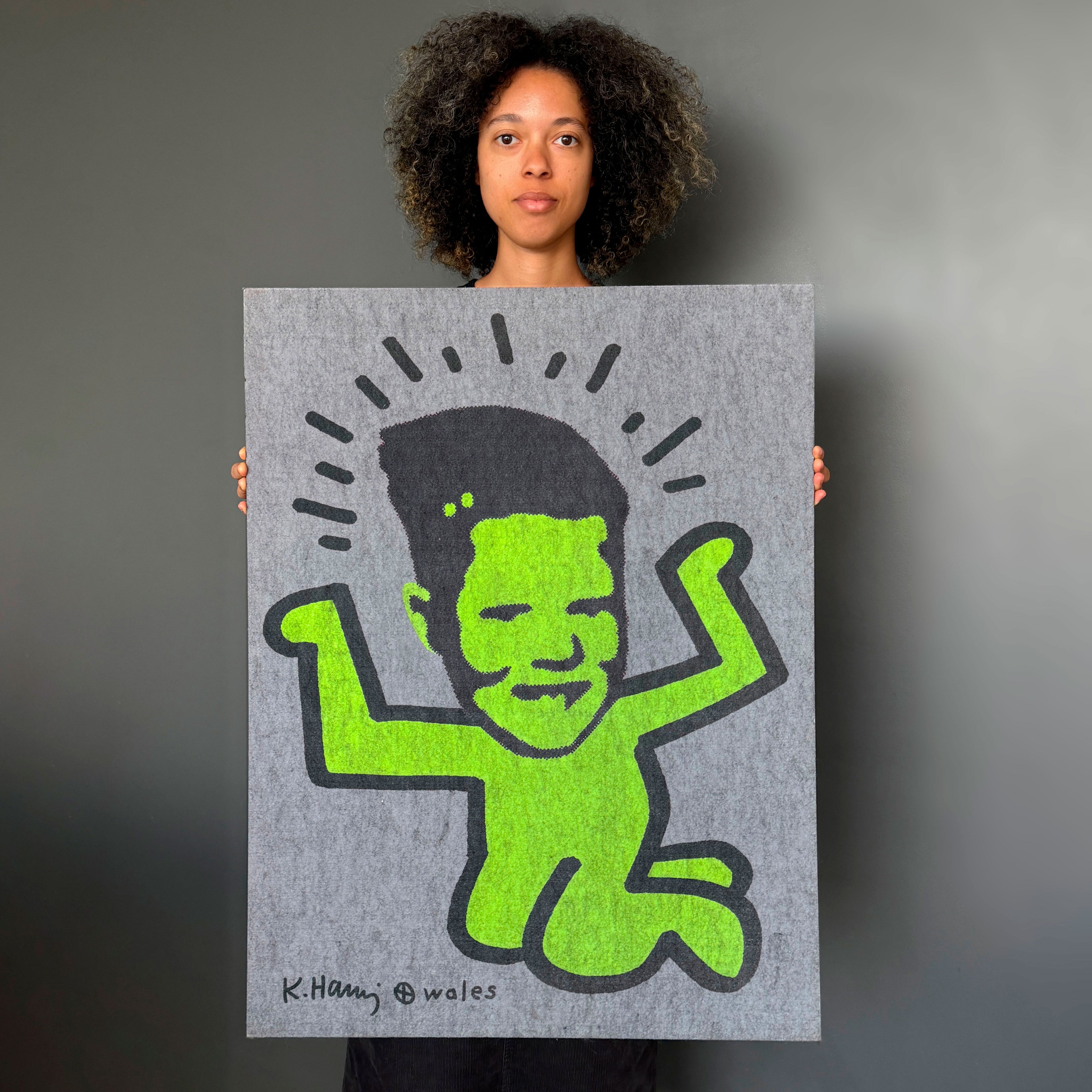 A person holds up a large square art print featuring a stylized green figure with black hair and black outlines on a gray background, against a dark gray wall.