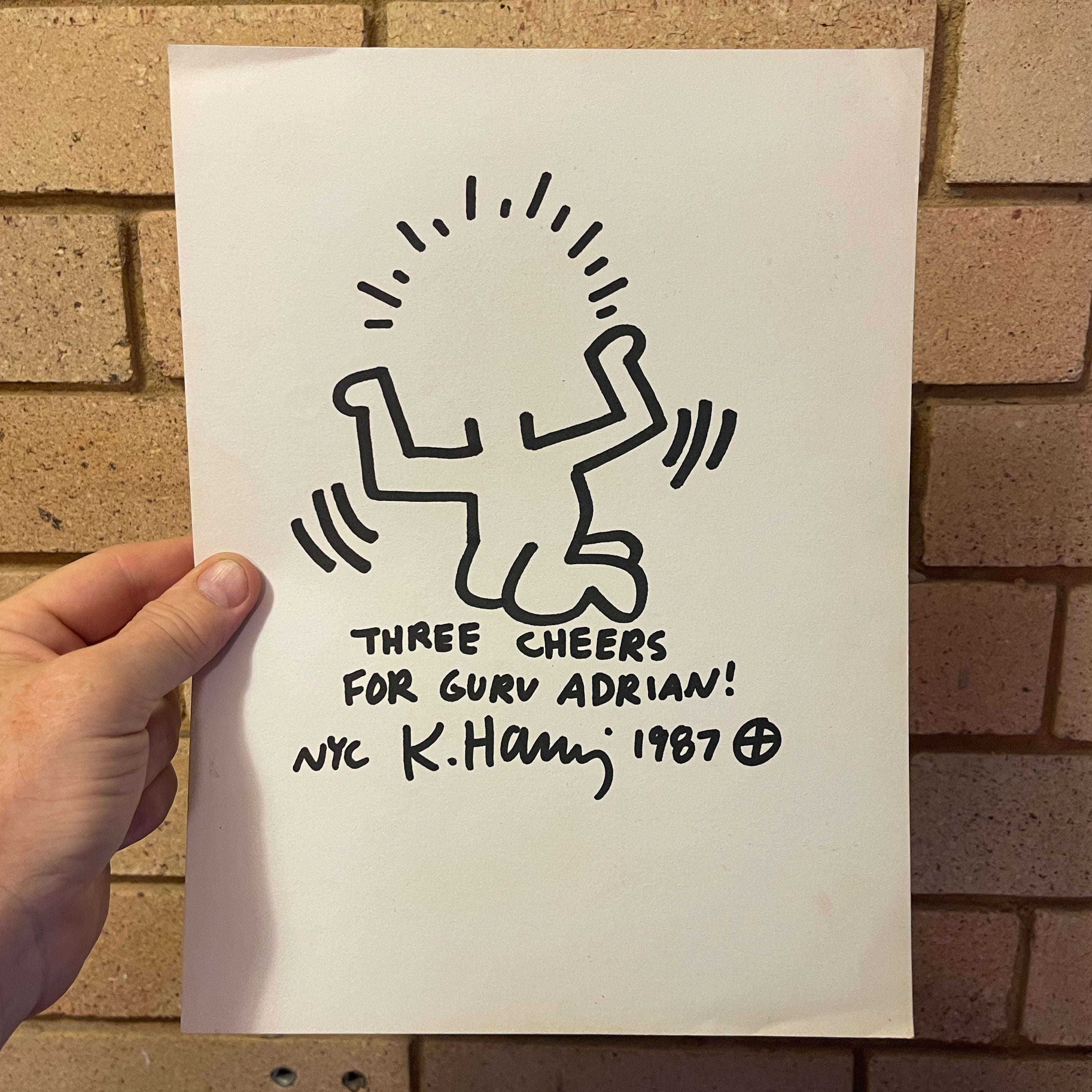 A hand holds a white art print featuring a black line drawing in the style of Keith Haring, with the text 'THREE CHEERS FOR GURU ADRIAN! NYC K. Haring 1987' against a tan brick wall background.