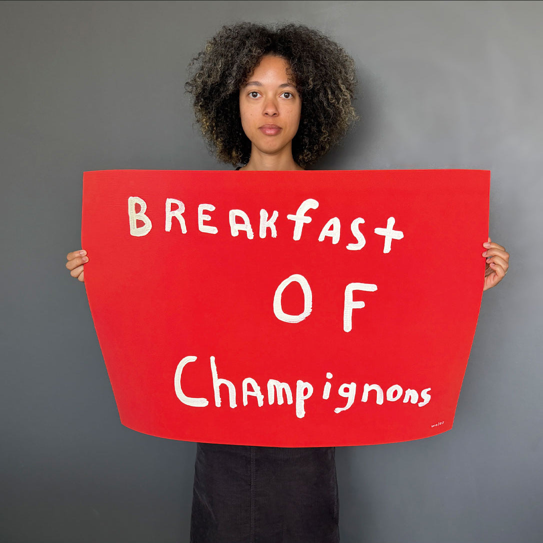 A person with curly dark hair holds a large red sign with white handwritten text reading 'Breakfast of Champignons' against a plain gray background.