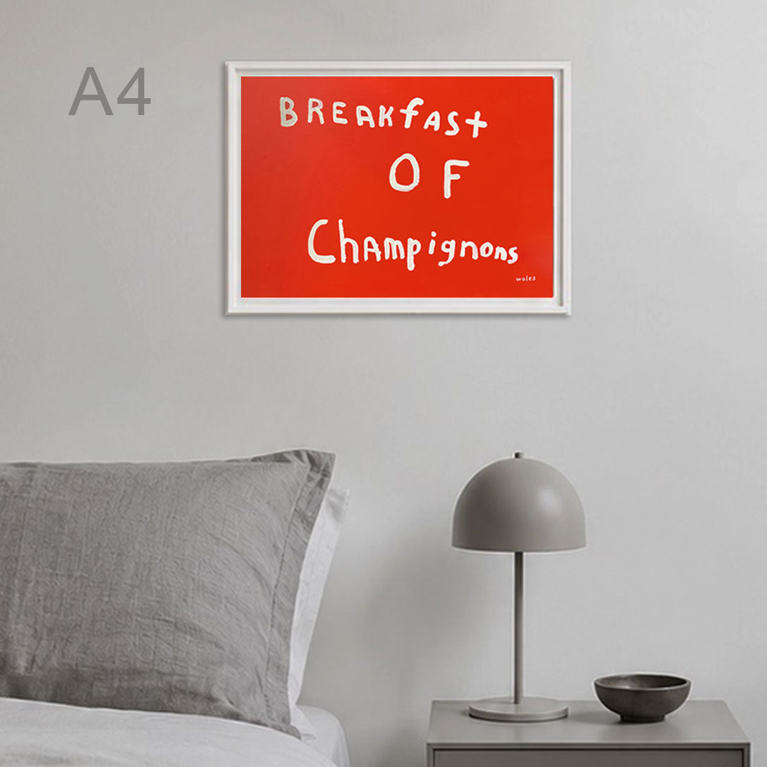 A framed wall art print with the phrase 'Breakfast of Champignons' in white handwritten text on a bright red background, shown hanging above a bed in a minimalist bedroom setting.