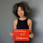A woman with curly dark hair wearing a black tank top holds a bright red sign that reads 'Breakfast of Champignons' against a plain gray background.