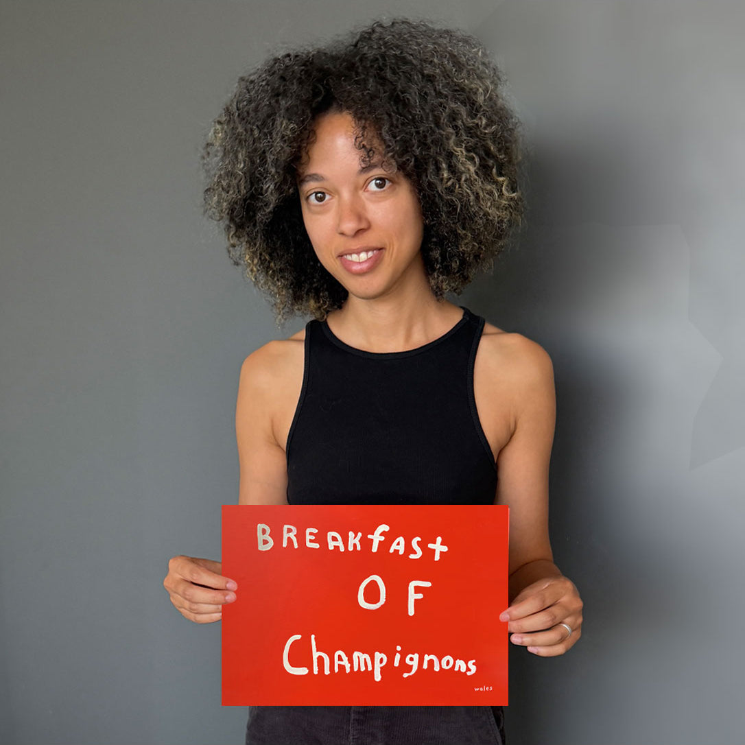 A woman with curly dark hair wearing a black tank top holds a bright red sign that reads 'Breakfast of Champignons' against a plain gray background.