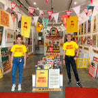 Two people standing in a retail store or art gallery hallway wearing matching bright yellow T-shirts that read 'DAVID'S ART SALE'. The space has colorful vintage-style posters, hanging sale flags, display tables, and a concrete floor with a red carpet runner.