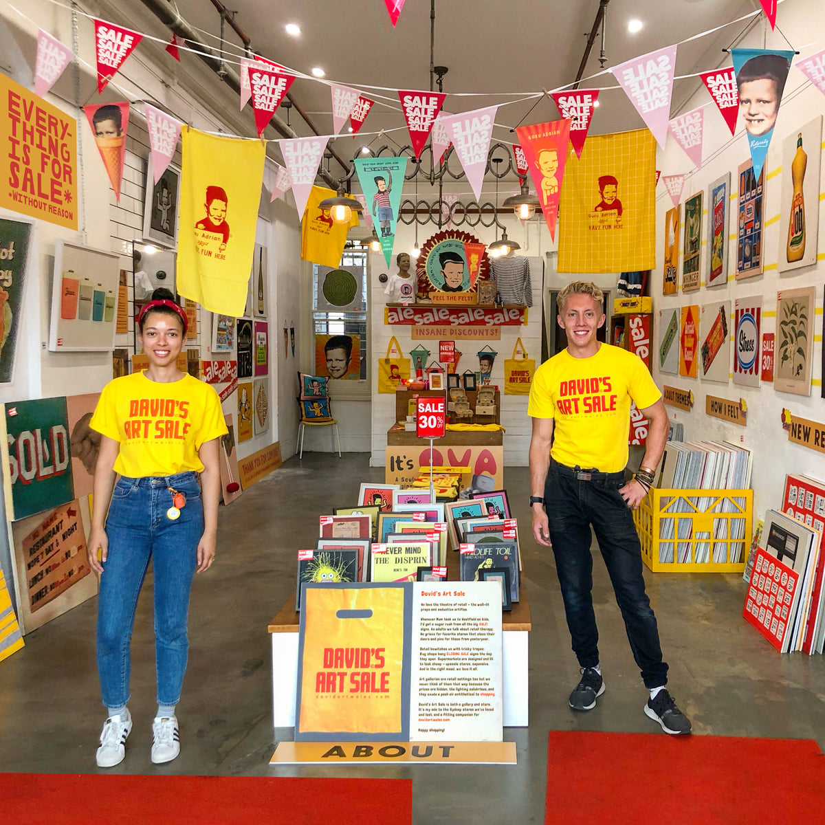 Two people standing in a retail store or art gallery hallway wearing matching bright yellow T-shirts that read 'DAVID'S ART SALE'. The space has colorful vintage-style posters, hanging sale flags, display tables, and a concrete floor with a red carpet runner.