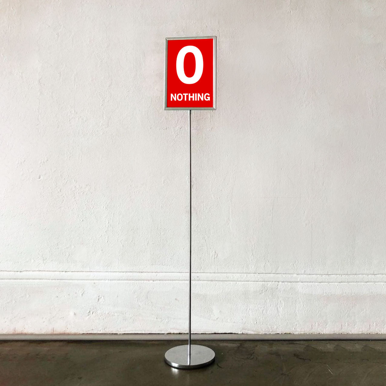 A freestanding metal sign on a thin pole with a round base, showing a red square sign that reads '0 NOTHING' in white text, set against a textured off-white wall and dark floor.