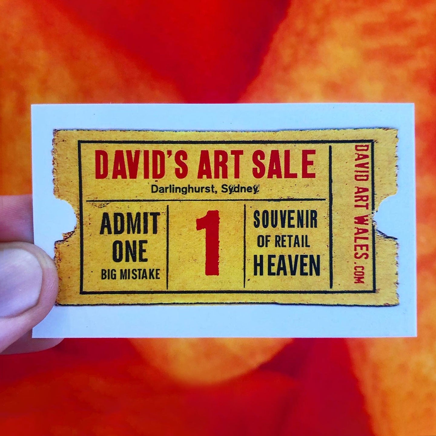 A hand holds a rectangular printed ticket or card featuring a vintage-style art sale design. The card has a yellow background with red and black text reading 'DAVID'S ART SALE,' 'Darlinghurst, Sydney,' 'ADMIT ONE BIG MISTAKE,' '1,' 'SOUVENIR OF RETAIL HEAVEN,' and 'DAVID ART WALES.