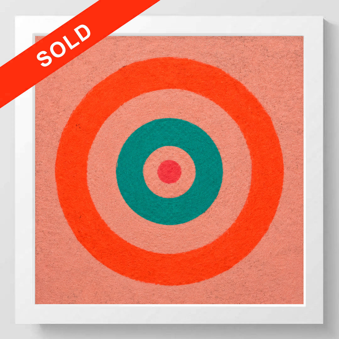 A framed square art print featuring concentric target-like circles in coral orange, teal, and pink on a peach background, shown against a light gray wall. A red diagonal banner across the top left says SOLD in white text.