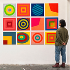 A person stands in a gallery or studio looking at a 3x4 grid of colorful abstract square artworks mounted on a white wall. The paintings feature bold geometric shapes, concentric circles, and color blocks in red, yellow, blue, green, orange, and pink.