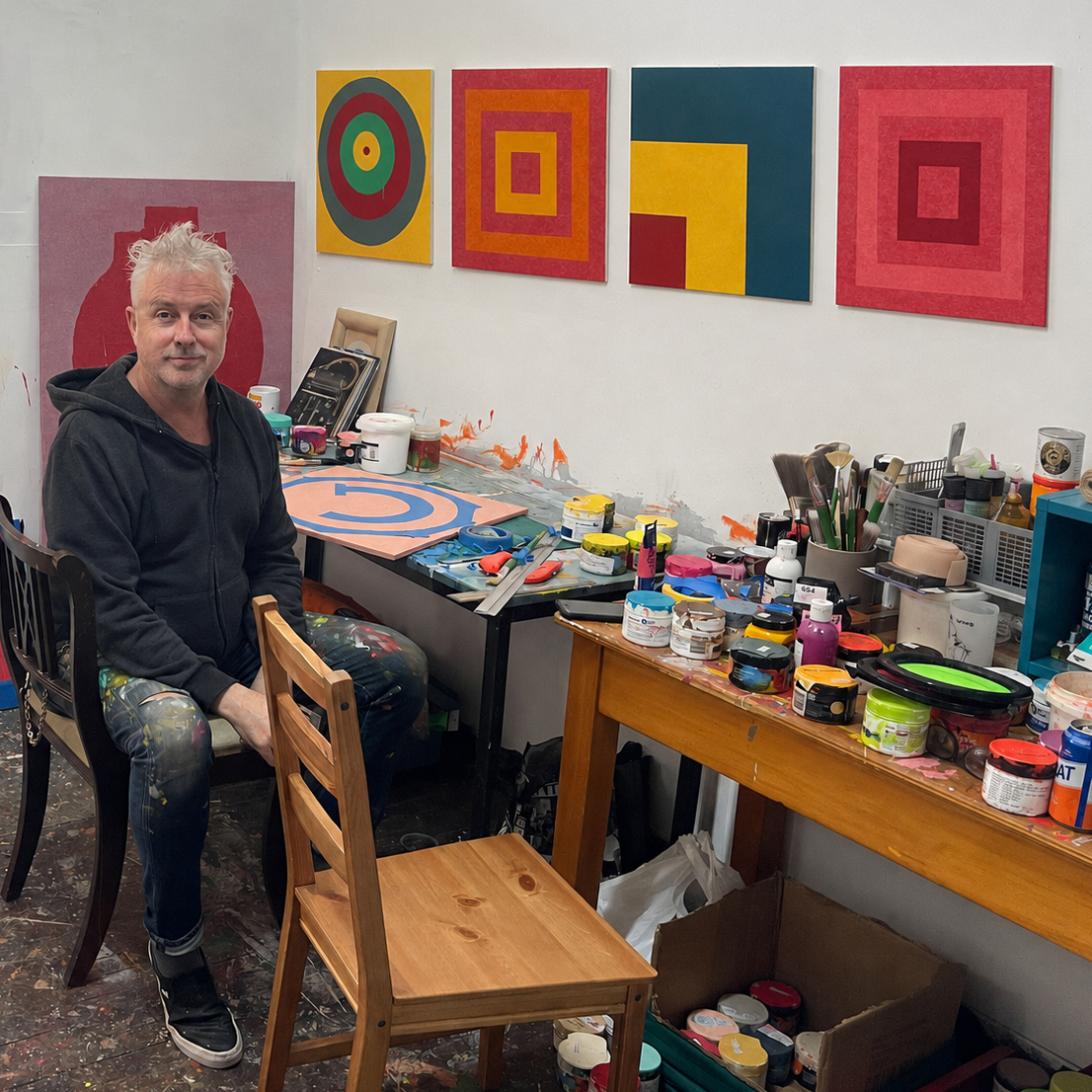 A man sitting in a paint-splattered artist studio with colorful geometric paintings hanging on the wall behind him, a wooden chair in the foreground, and worktables covered with paint cans and brushes.