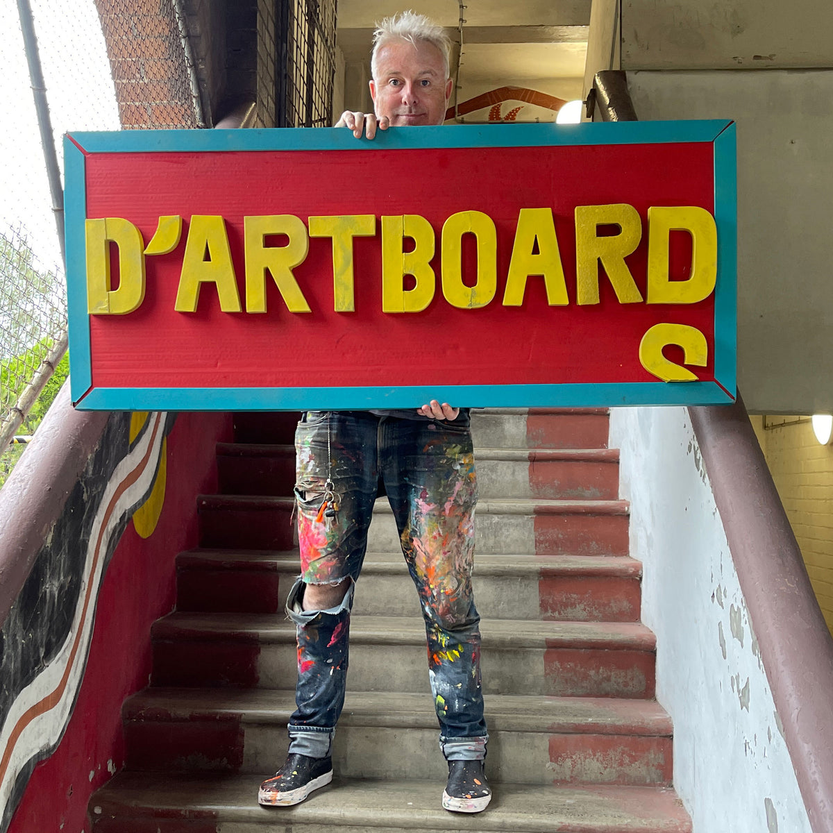A man with short blond hair stands on painted stairs holding a large rectangular sign that reads “D'ARTBOARD” in yellow letters on a red background with a turquoise border.