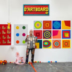 An artist stands in a gallery or studio space in front of a white wall, holding two small colorful objects. The wall is covered in colorful abstract and geometric paintings, including a red sign reading 'D'ARTBOARD' at the top.