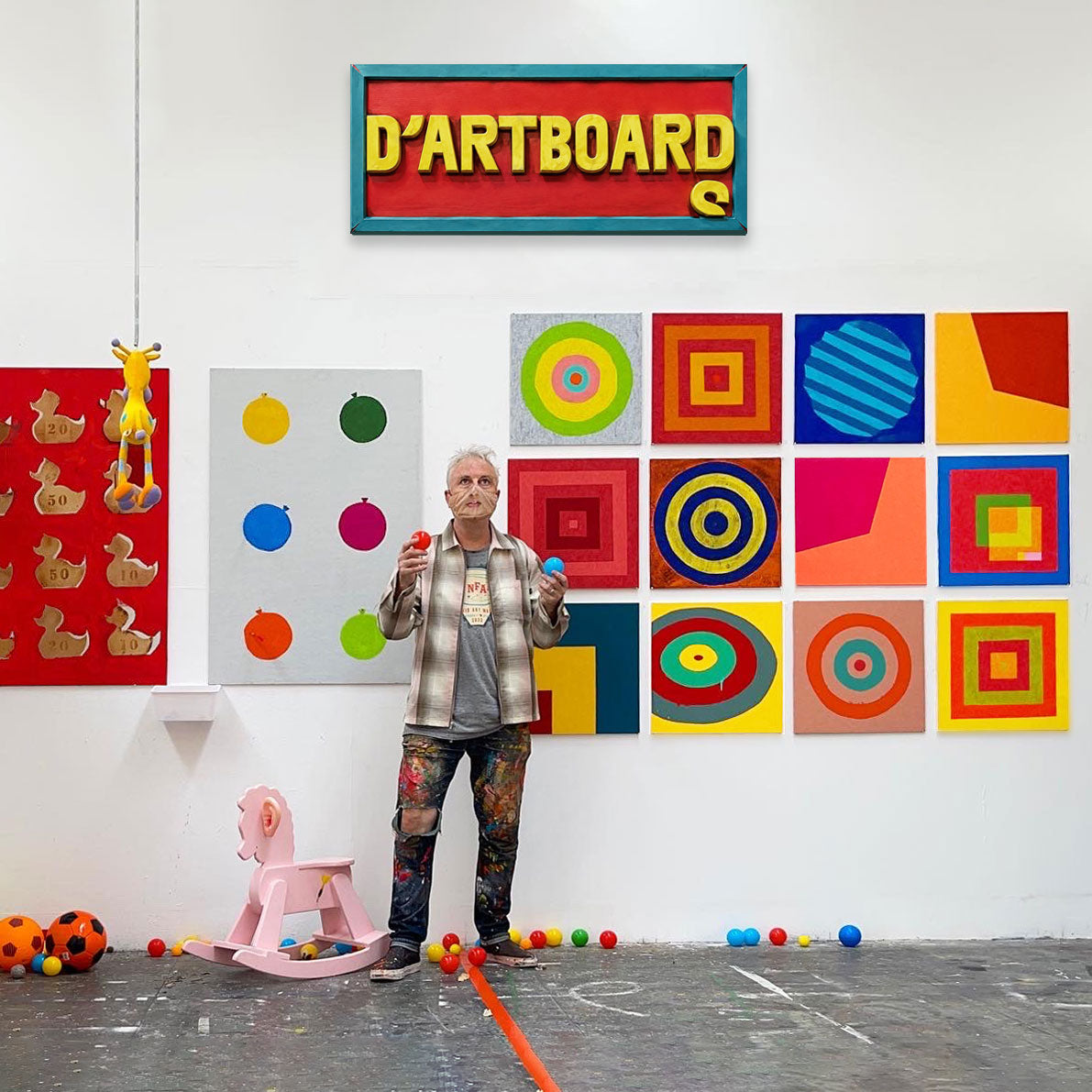 An artist stands in a gallery or studio space in front of a white wall, holding two small colorful objects. The wall is covered in colorful abstract and geometric paintings, including a red sign reading 'D'ARTBOARD' at the top.