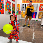 A toddler in a red dinosaur-patterned hooded rain suit holds a large yellow tennis ball in a gallery space with colorful abstract paintings on the walls. An adult in a yellow and black jacket stands in the background holding a drink.