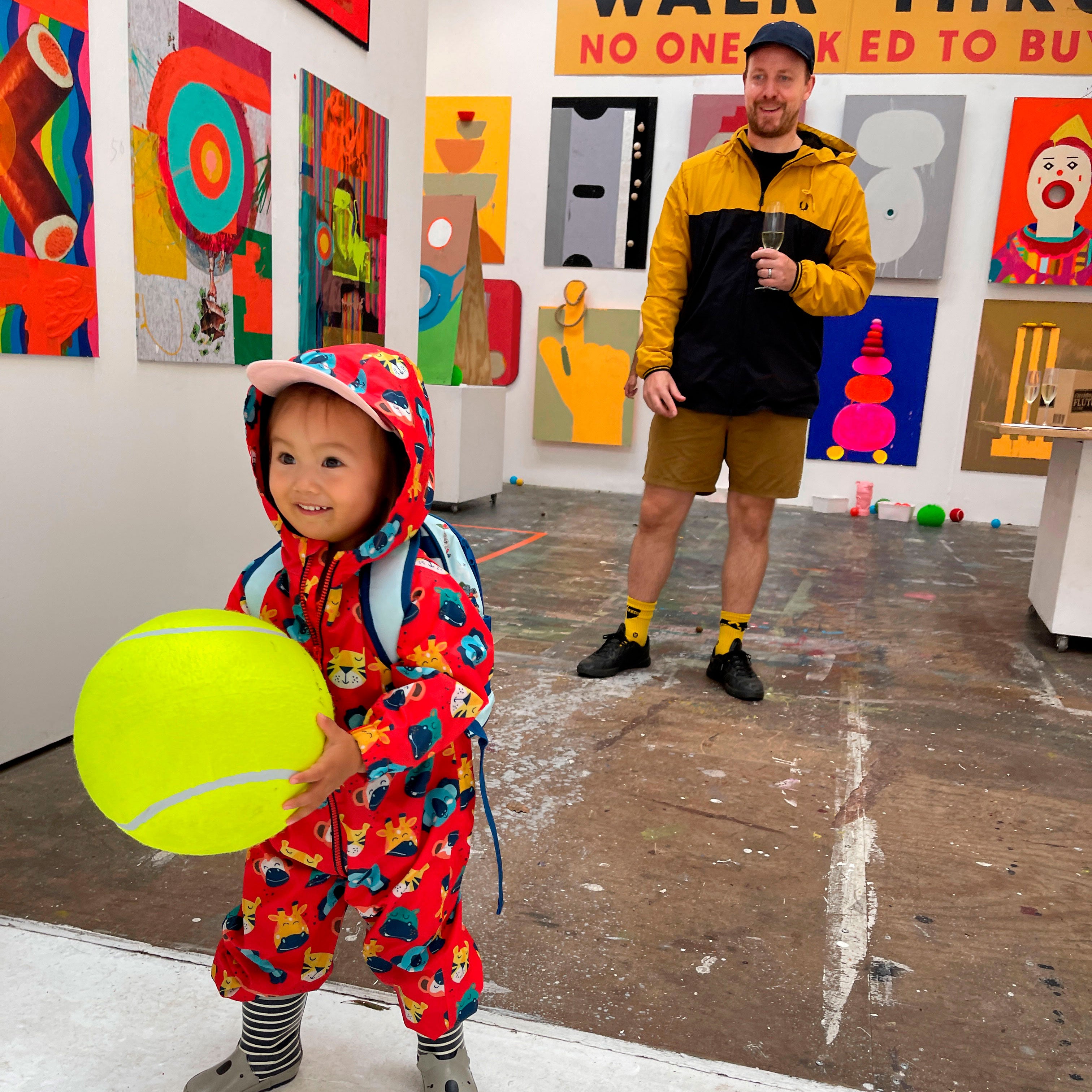 A toddler in a red dinosaur-patterned hooded rain suit holds a large yellow tennis ball in a gallery space with colorful abstract paintings on the walls. An adult in a yellow and black jacket stands in the background holding a drink.
