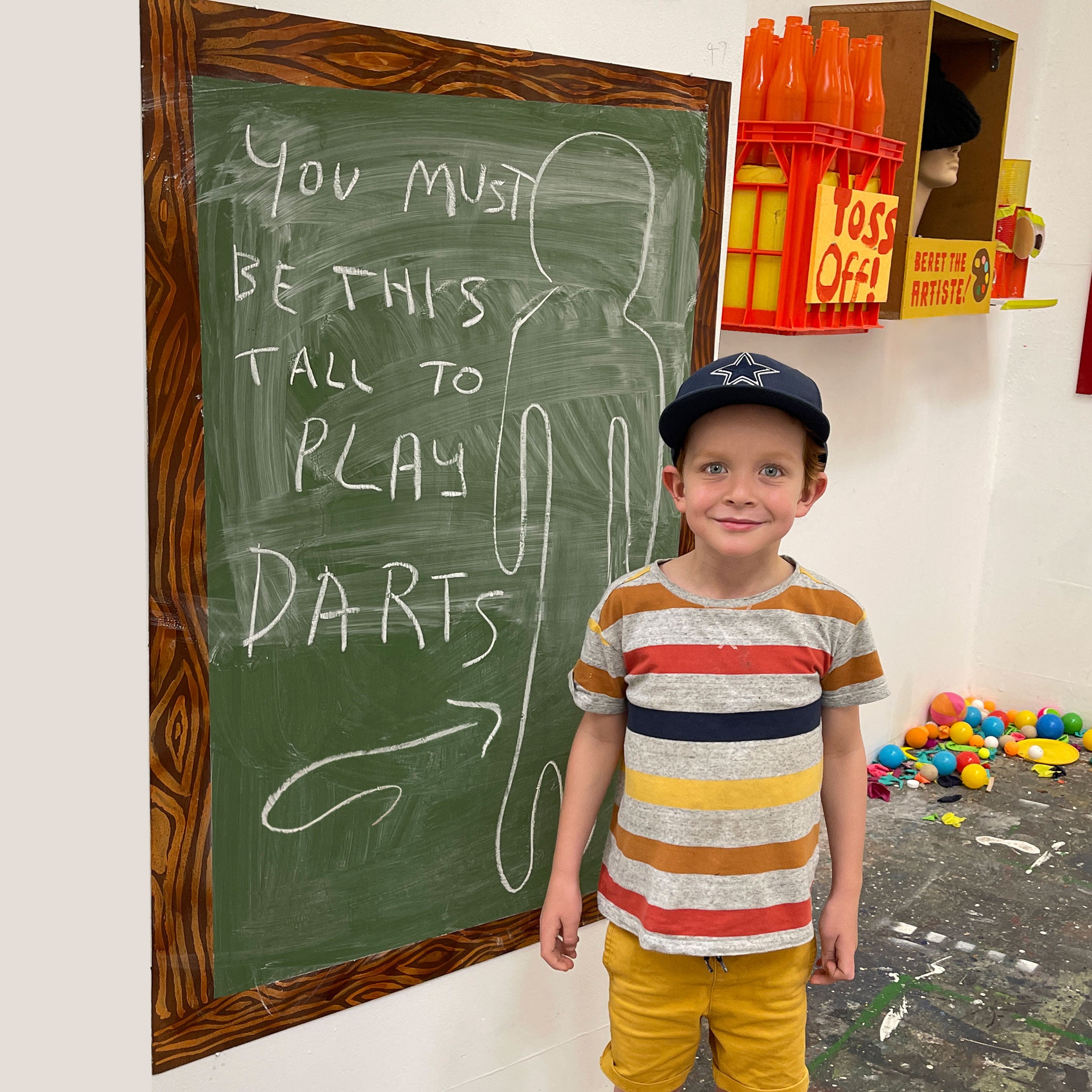 A young boy wearing a navy blue cap and a colorful horizontally striped t-shirt stands in front of a chalkboard that reads 'YOU MUST BE THIS TALL TO PLAY DARTS' in a playful indoor setting.