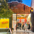 A group of people standing in the doorway of a retail store at dusk, with a large yellow 'FUNFAIR' sign on the left and a 'WALK THRU NO ONE ASKED TO BUY' sign above the entrance. The building has a rustic brown wooden facade and a blue evening sky.