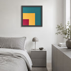 A framed abstract geometric wall art print hangs above a bed in a modern, neutral-toned bedroom. The artwork features a teal background with yellow and magenta geometric blocks, enclosed in a dark gray frame.