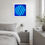 A framed abstract art print featuring a blue circular design with diagonal stripes, displayed on a light gray wall above a modern gray bed in a minimalist bedroom setting.