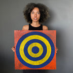 A woman with curly dark hair holds a square canvas painting featuring concentric circles in blue and yellow on a textured orange-red background, posed against a solid gray studio backdrop.